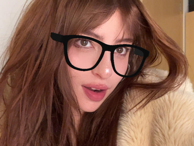 AngelinaNoAngel has brown hair, black glasses, and is looking at the camera with her mouth slightly open. She's wearing a furry coat. Her tits are not visible.