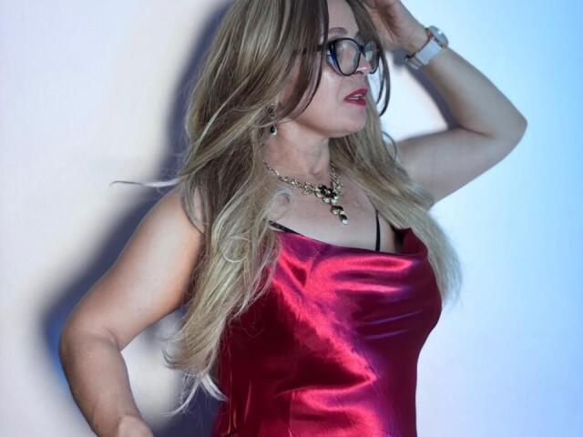 SamantaMature69 with long blonde hair, glasses, red satin dress, necklace, hand on head, looking to the side, blue wristwatch, red lipstick, standing against a light background.
