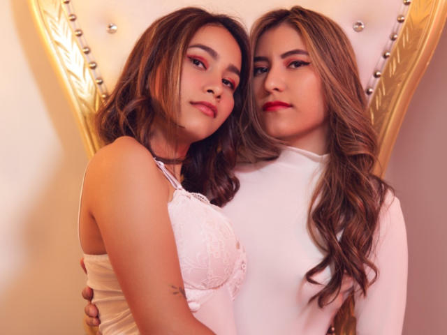 SamantaAbby con cabello castaño, usando una blusa de encaje, y SamantaAbby con cabello rubio, usando una blusa blanca, ambas con lápiz labial rojo, con los brazos alrededor una de la otra, mirando a la cámara..