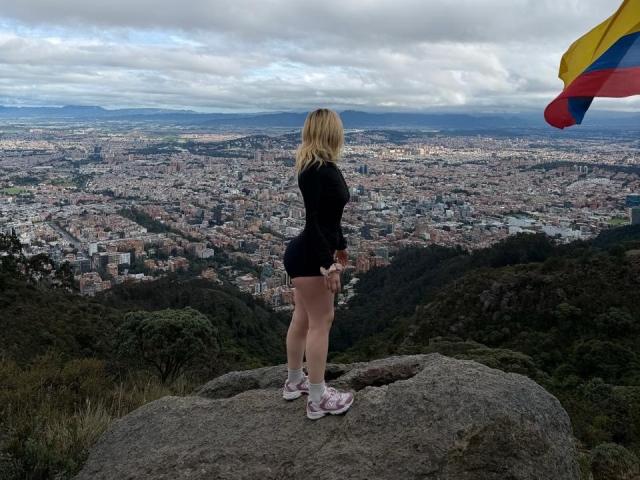 SammyViolet with blonde hair, black long-sleeve top, and shorts, standing on a rock, looking at a city with a colorful flag on the right. She has a cute ass.