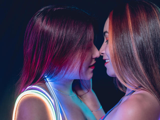 Samanta e Abby, com cabelos ruivos e castanhos, luzes neon azuis e rosas, uma de frente para a outra, com o nariz quase tocando, sorrindo, vestindo blusas sem mangas, exibindo seios pequenos, em um ambiente escuro..