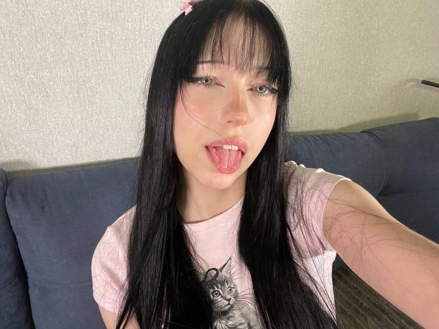 AmaiRin, avec ses cheveux noirs, son serre-tête rose et son t-shirt rose, tirant la langue, montrant son petit nez et ses lèvres roses, avec un mignon motif de chat gris..