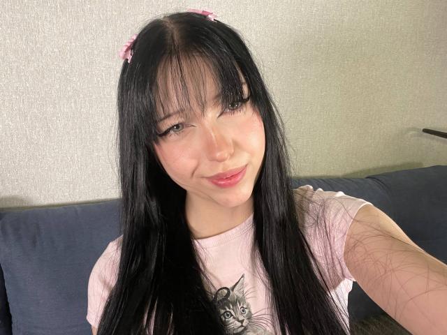 AmaiRin avec de longs cheveux noirs, des pinces à cheveux en forme de fleurs roses, une chemise rose, souriante, mignonne, petite poitrine, peau claire, yeux marrons, tatouage sur le bras, assise sur un canapé bleu, selfie..