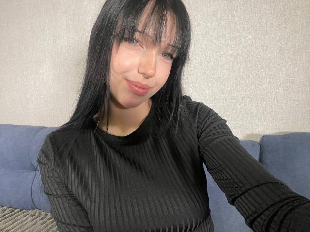 AmaiRin portant une chemise noire à rayures, avec de longs cheveux noirs lisses, et une bouche mignonne, souriant légèrement, assise sur un canapé bleu, montrant de petites seins galbés, et un visage adorable..