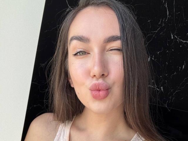 AraiaSheri has pouty pink lips, one green eye winking, long brown hair, and a fair complexion, wearing a white tank top, against a black marble-like background.