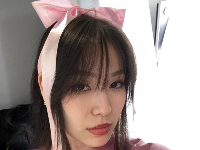 CatherinaKicker with pink bow headband, dark straight hair, and red lipstick, looking into camera. She has light skin, small nose, and subtle makeup. Her lips are slightly parted.