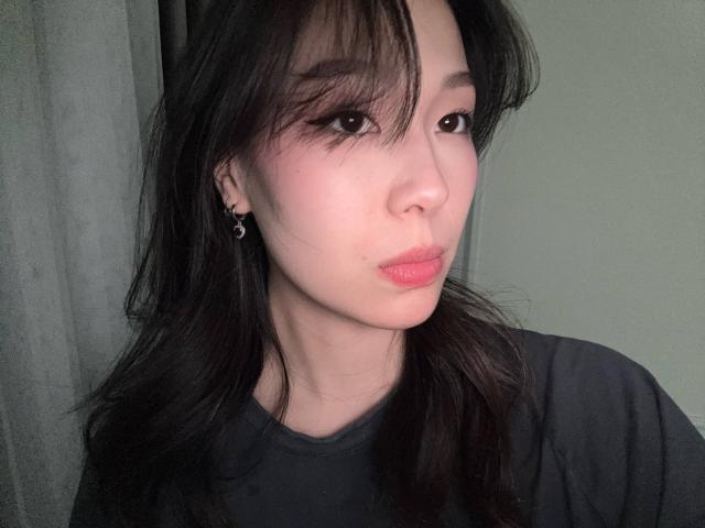 CatherinaKicker has pale skin, dark straight hair, pink lipstick, and small earrings. She's pouting with her lips slightly up. She's wearing a black shirt.