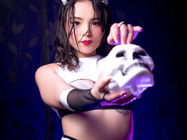 ViolettaMidnight holding a skull with her right hand, wearing a white crop top, black mesh arm sleeves, and a maid headpiece, with wet black hair and red lipstick.