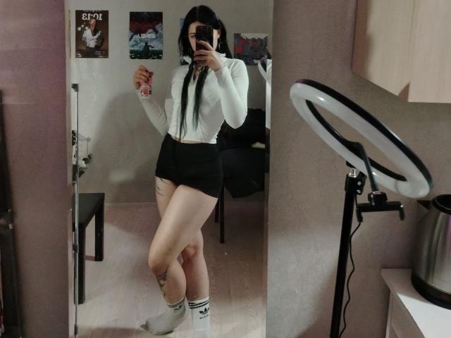 JanetWiedemann with long black hair, white long-sleeve shirt, black shorts, white socks, holding phone, mirror selfie, ring light on right, visible thigh tattoo, standing in room.
