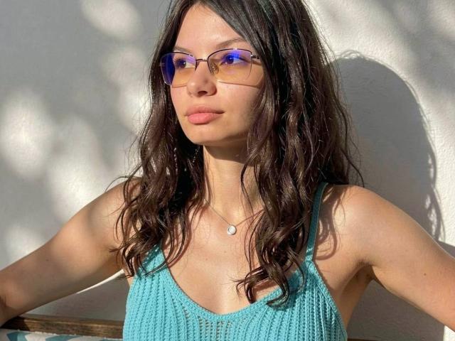 VikkiRi with wavy dark brown hair, wearing a turquoise knit top, blue reflective sunglasses, and a small necklace, standing in sunlight, showing her bare tits and ass.