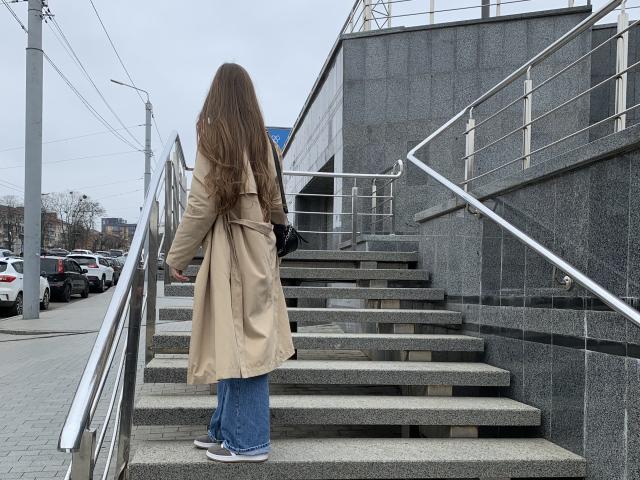 LexiLore, back to the camera, wearing a tan coat, blue jeans, and white sneakers, climbing grey stone stairs with metal railings, holding a black purse.