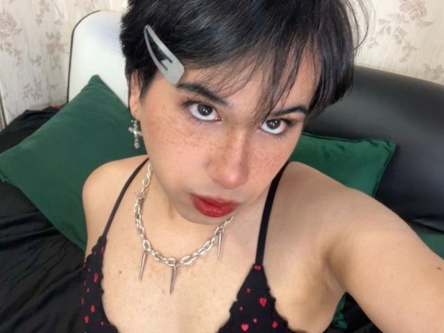 FemCupid with short black hair, gray hairclip, red lipstick, freckles, chain necklace with spikes, black lace top, green pillows, and black bed, looking up at camera.