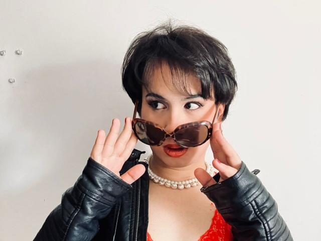 FemCupid con cabello negro corto, vistiendo una chaqueta de cuero negro, una blusa de encaje rojo, un collar de perlas y grandes gafas de sol de color marrón, con las manos ajustando las gafas, con expresión de sorpresa..