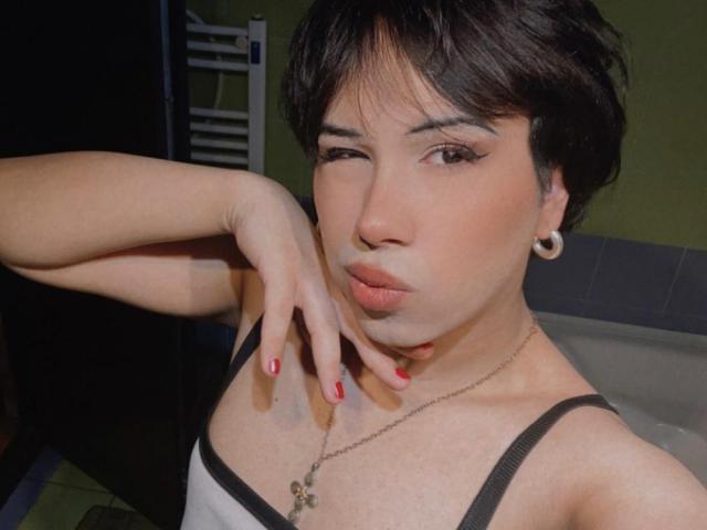 FemCupid with short black hair, wearing a white tank top, has red nails, a cross necklace, and black ear gauges, puckering her lips, with a hand near her chin.