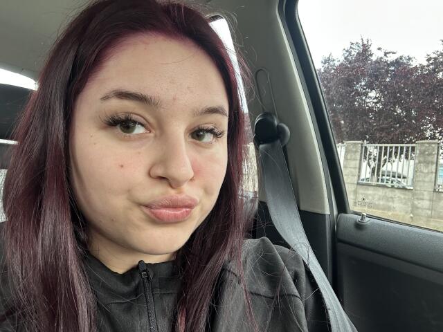 LucieRed a la peau pâle, les yeux verts et les cheveux avec des pointes violettes, et elle fait la moue avec ses lèvres roses, assise dans une voiture attachée avec une ceinture de sécurité. Elle porte une chemise noire à fermeture éclair..
