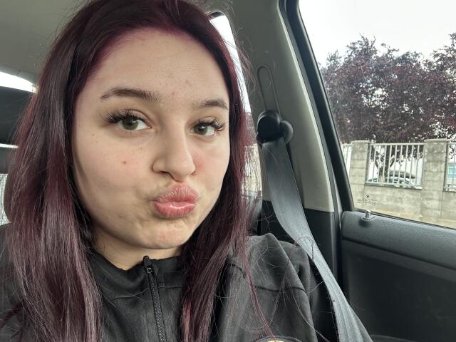 LucieRed a la peau pâle, les yeux verts, et un rouge à lèvres rose-violet, ses lèvres brillantes étant légèrement retroussées. Elle est assise dans une voiture, a les cheveux foncés et porte une veste noire à fermeture éclair..