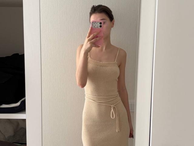 DanicaStuer with small tits in a tight, beige, ribbed dress, holding a pink phone, mirror selfie, in a minimalistic room. She's wearing a tied waist, standing straight.