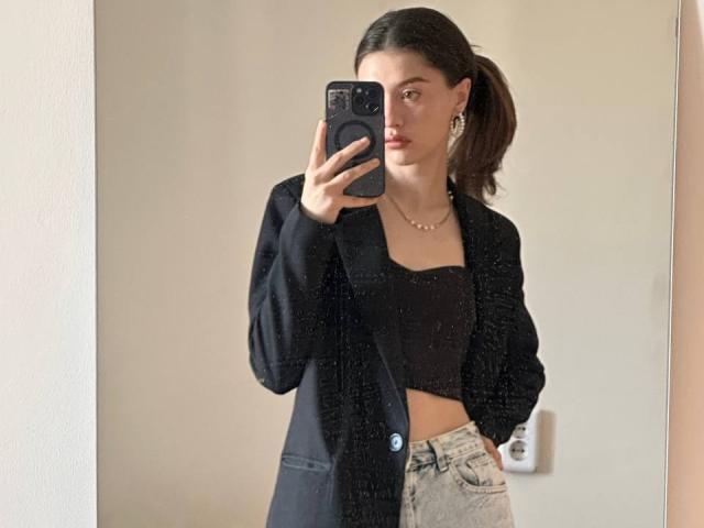 DanicaStuer with dark hair in a ponytail, wearing a black jacket, black crop top, and light jeans, taking a mirror selfie with a serious expression, showing her tits.