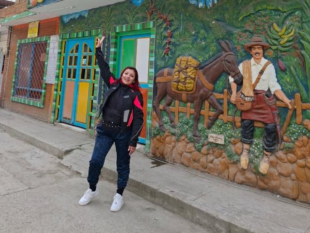 LizBonnet stands with arm raised, wearing a black jacket with red sleeves, blue jeans, and white sneakers, next to a painted donkey and cowboy on a colorful wall.