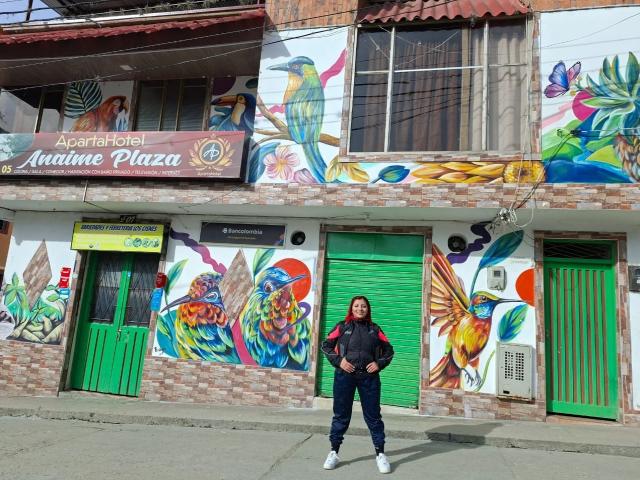 LizBonnet stands in front of a colorful building with peacock and butterfly graffiti, wearing a black jacket, blue pants, and white sneakers, with a green door behind her.