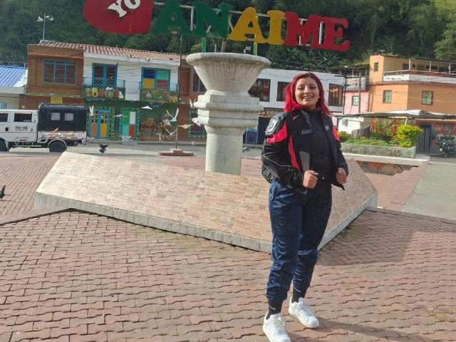 LizBonnet stands in a plaza wearing a black and red jacket, navy pants, and white sneakers. She has red hair, a medium build, and a happy expression.