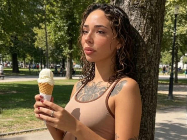 SilvanaXOwen with curly brown hair, tan tank top, holding a vanilla ice cream cone, showing off her tits and tattoos, standing against a tree in a park.