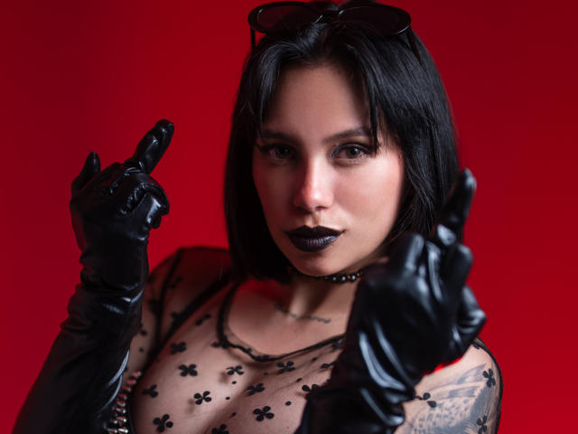 LilithDeveraux avec une coupe de cheveux courte noire, du rouge à lèvres foncé, des gants noirs, un haut transparent à motifs floraux, pointant du doigt avec sa main droite, des lunettes de soleil noires sur la tête, un fond rouge, un tatouage sur le bras droit..