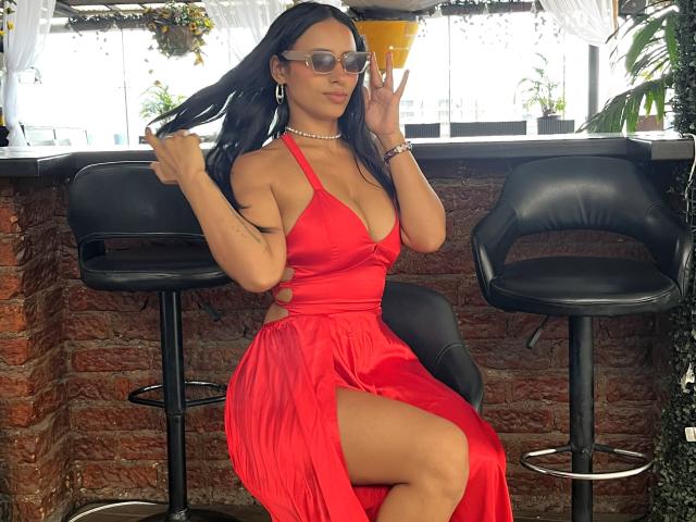 Nicol Santos indossa un abito rosso con una profonda spaccatura che rivela la coscia, è seduta su uno sgabello nero, si tiene i capelli, indossa occhiali da sole e una collana di perle..
