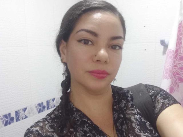 HypnoticBella has a cute face with pink lipstick, black braided hair, and is wearing a black lace top, looking at the camera with a slight smile.