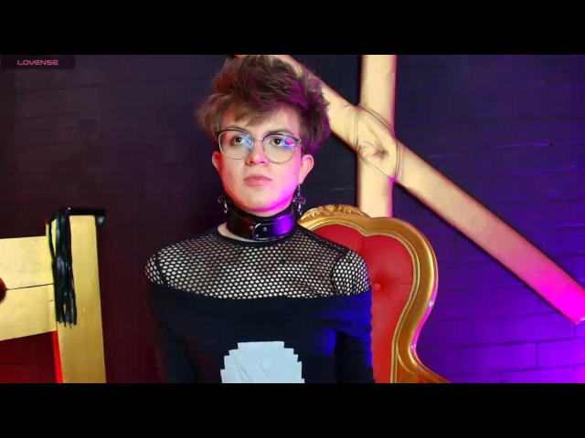 DionnySian with short brown hair, glasses, and a black mesh top with a white pixel design, wearing a black collar, standing in front of a red and gold chair.