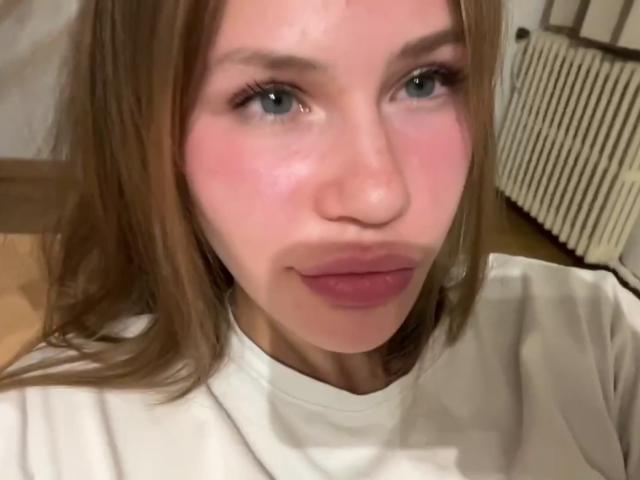 DedeUrtiaga's face with pink cheeks, blue eyes, and brown hair in a white shirt, pouting pink lips, slightly flushed, looking to the side.