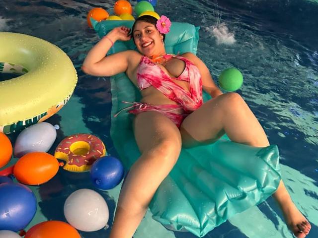 CristalZafiro, uma mulher corpulenta com seios grandes e coxas grossas, está deitada em um brinquedo inflável verde, vestindo um biquíni rosa tie-dye, cercada por balões coloridos em uma piscina..