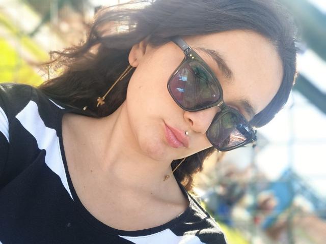 NataliaRivas with dark sunglasses, black-and-white striped shirt, and a nose piercing, smiling slightly. She has wavy brown hair and gold earrings. Sunlight reflects off her glasses.