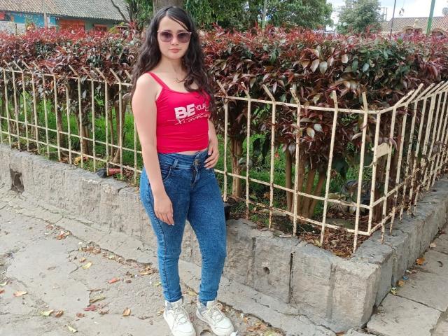 NataliaRivas with medium tits in a red tank top, blue jeans, white sneakers, and pink sunglasses, standing in front of a fenced garden with red-leaved bushes.
