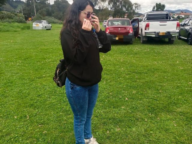 NataliaRivas stands on grass, wearing a black hoodie, blue jeans, and white sneakers, holding a phone to her ear, with a black backpack and long wavy hair.