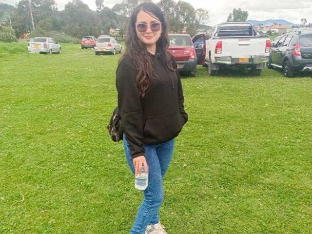 NataliaRivas has long dark hair, wearing sunglasses, a black hoodie, blue jeans, and white sneakers, holding a water bottle, standing on green grass with parked cars.