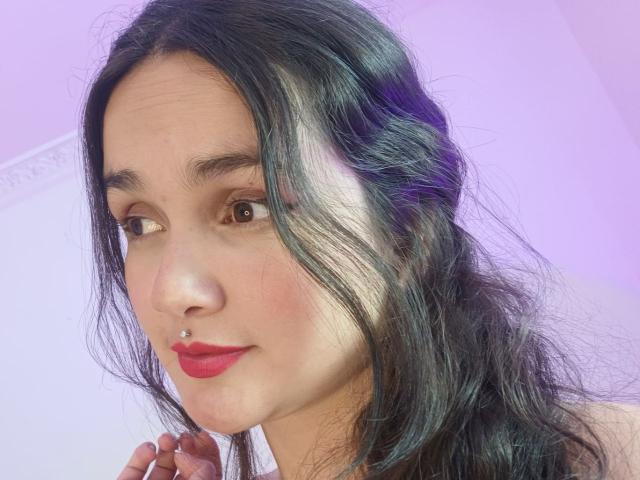 NataliaRivas has long black hair in a loose side braid, a silver nose piercing, and red lipstick. She's looking to the side with brown eyes and a slight smile.