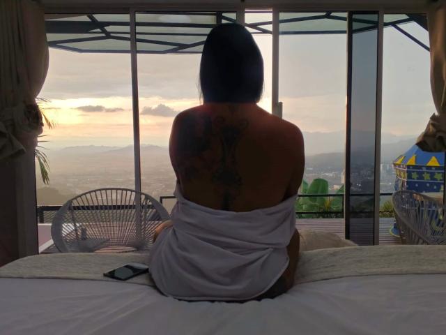 JessicaFiore, back to camera, bare-assed, with white towel covering her ass, black bob haircut, large tattoo on her back, sitting on bed, sunset view, phone on bed.