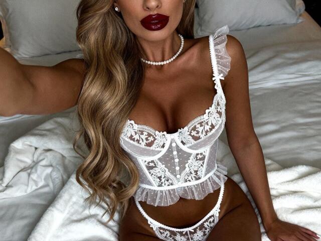 ElzaKomx with wavy blonde hair, wearing a white lace corset and matching panties, showing her tits, a pearl necklace, and dark red lipstick, sitting on a bed.