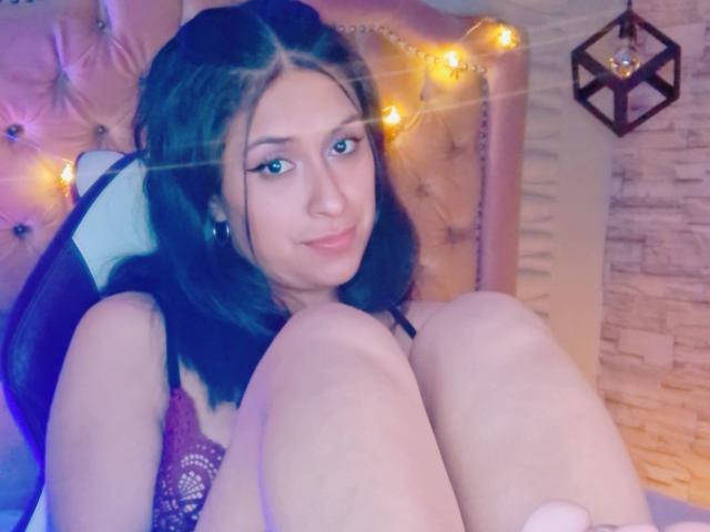 RoxanneHan, com cabelo castanho escuro, usando um sutiã de renda roxo, sentada com os joelhos dobrados, com um sorriso adorável, seios pequenos, olhos azuis e luzes de fada amarelas quentes ao fundo..