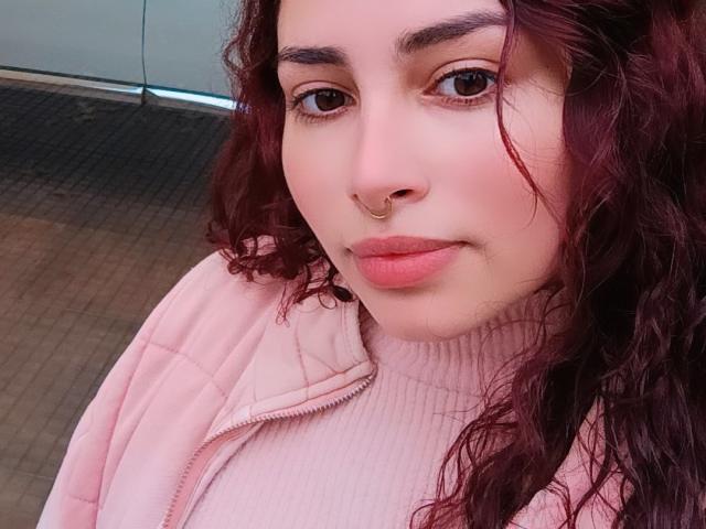 SweetSolei69 ha i capelli rossi ricci, un anello dorato al naso e rossetto rosa, indossa un maglione a coste rosa e una giacca abbinata, con un'espressione leggermente corrucciata..