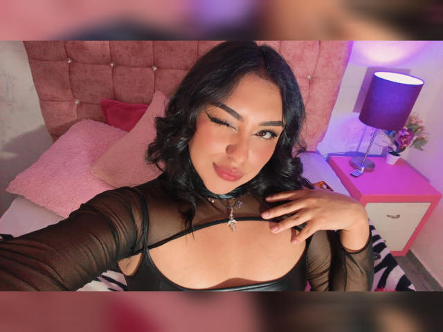 SyraPalmer, avec un clin d'œil, des cheveux noirs bouclés, un haut noir transparent, une décolleté visible, des coussins roses, une lampe rose, une parure de lit zébrée. Sa main touche sa poitrine, un collier est visible..