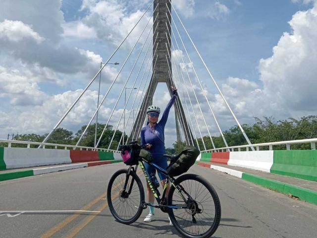 QueenScarletSecret, with a blue spandex suit and helmet, stands on a bridge, arm raised, holding a black bike with a pink water bottle and black bag.