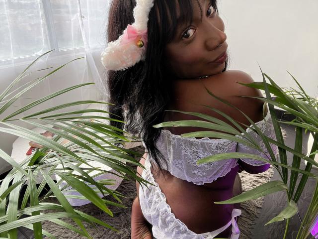 WarisEllen with dark skin, long black hair, wearing white lace off-shoulder lingerie and pink bunny ears, looking back, showing her bare ass, standing in front of green plants.