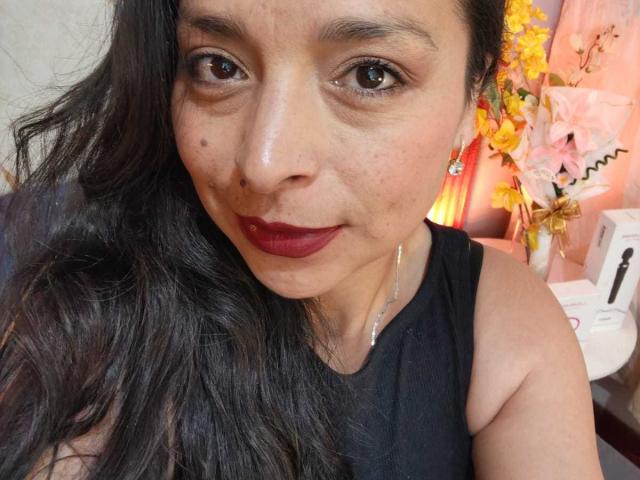StangelarRivera, met lang zwart haar, bruine ogen, donkerrode lippenstift, een klein beauty markje op de wang, draagt een zwart topje zonder mouwen, oorbellen en een subtiele ketting, en glimlacht naar de camera..