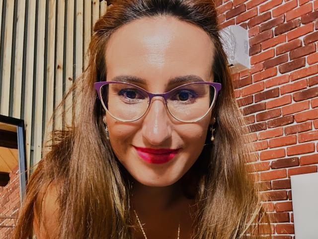 LysAddiction with purple glasses, red lipstick, and long brown hair, smiling at the camera, her tits slightly visible under a shirt, against a brick wall.
