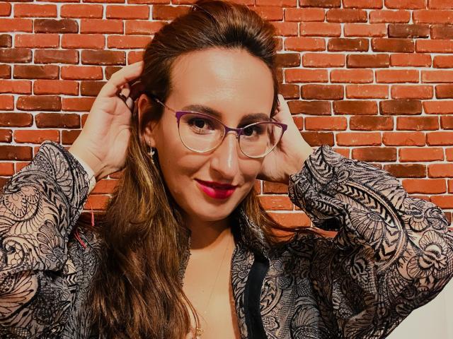 LysAddiction with glasses, red lipstick, and brown hair, hands in her hair, wearing a black and white patterned shirt, smiling. She's got a cute ass.