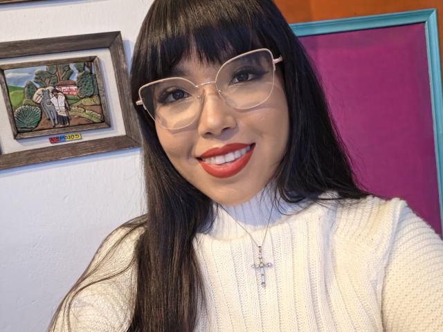 Heyly, met lang zwart haar, een bril en een witte coltrui, lachend met rode lippenstift, die haar schattige gezicht en haar kruisbedeltje laat zien, en die er sexy en aantrekkelijk uitziet..