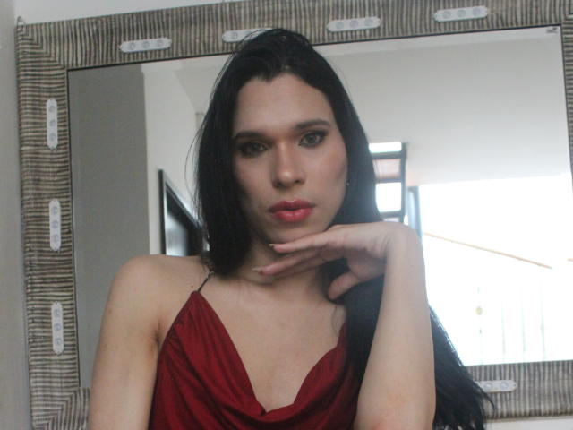 Jade Oconner mit langem, schwarzem Haar, einem roten Kleid und vollen Lippen, die ihr Kinn in die Hand legt, wobei sie ihre Brüste zur Schau stellt und im Spiegel in einen verführerischen Blick..
