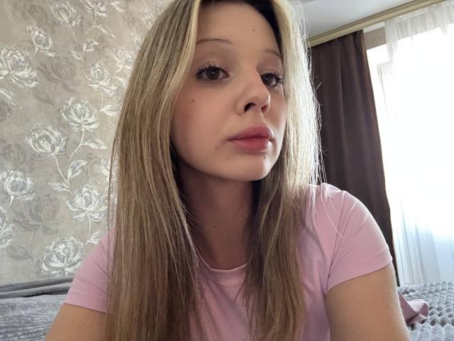 MerrieHamad heeft lang blond haar, draagt een roze shirt, heeft een neutrale uitdrukking, lichtroze lippen die iets open zijn, en een zichtbare neus, in een goed verlichte kamer met bruine gordijnen..